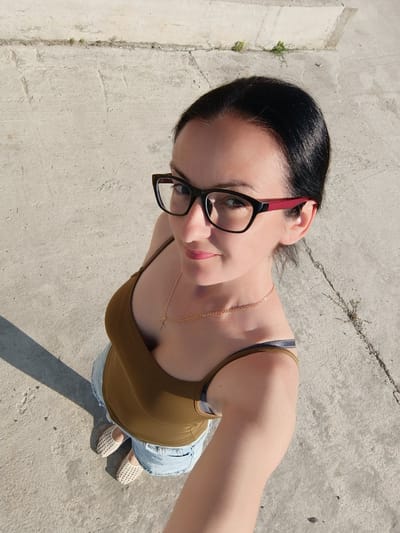 This is a selfie taken by a woman with dark hair and glasses, wearing a green tank top, denim shorts, and light-colored woven shoes. She is standing outdoors on a concrete surface, likely in Târgu Jiu, Romania. The lighting suggests it is daytime, possibly late afternoon due to the shadows cast by her and the surrounding structures. The concrete appears weathered and there are patches of sparse vegetation growing in cracks, indicating an outdoor environment. The woman is looking up towards the camera, and her expression is neutral to slightly smiling. A thin gold chain with a small cross pendant is visible around her neck. The background consists of the concrete ground and a raised concrete edge or wall. There is no visible text in the image.