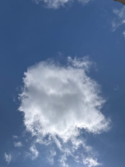 A photograph of a cloudy sky with a large, puffy cloud dominating the center of the frame. The sky is a clear, deep blue. The cloud is white and appears to be illuminated by sunlight, casting subtle shadows within its mass. Small wisps of thinner clouds are scattered around the larger cloud, also against the blue sky. The image appears to be taken from a ground-level perspective looking upwards. No people, structures, or specific geographical landmarks are visible, making it impossible to determine a specific location within Татищев Погост, Russia, or any city.