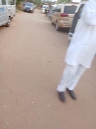 The image displays an unpaved, reddish-brown dirt road in Orie Uratta, Nigeria, under daylight. In the right foreground, a person is visible from the mid-torso downwards, wearing a white top, white trousers, and dark shoes, with significant motion blur indicating movement. On the left side of the road, a light-colored minivan is partially visible. Further down the road, a dark-colored SUV is present. On the right side, extending into the background, a line of various vehicles is observed, including a gold or beige SUV and other indistinct cars and motorcycles. The background features blurred indications of vegetation or distant structures.
