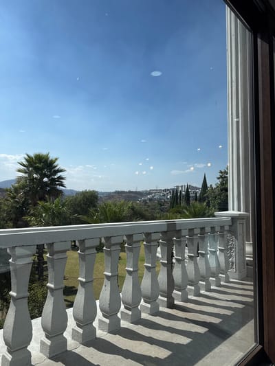 The image depicts an exterior view from behind a large window or glass door, showcasing a residential landscape in Atizapán de Zaragoza, Mexico. In the immediate foreground, visible through the glass, is a white, classical-style concrete balustrade serving as a balcony railing, casting sharp shadows onto the concrete balcony floor. To the far right, a segment of a fluted white column, part of the building's structure, is visible through the glass. The sky overhead is clear blue with minimal cloud cover, and reflections of interior circular recessed lights are visible on the glass surface.

Beyond the balcony, the midground features a verdant landscape dominated by various green trees and shrubs, including prominent palm trees on the left and a well-maintained grassy area directly behind the balustrade. The background consists of rolling hills sparsely covered with vegetation. On these distant hills, residential development is visible, characterized by numerous white structures and some red rooftops, consistent with suburban architecture in the Atizapán de Zaragoza area. A cluster of tall, slender cypress trees is also distinguishable amidst the distant buildings. No individuals, specific actions, or interactions are present in the scene.