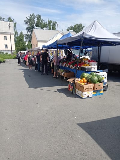 In the city of Degtyarsk, Russia, a busy outdoor market is depicted. A diverse group of people are browsing and shopping at various stalls set up under awnings. Several vendors are visible, attending to customers and displaying a variety of goods. Produce, such as watermelons, oranges, and other fruits and vegetables, are prominently featured in boxes and on tables. Clothing items are also visible on display at some stalls. The market appears to be set up on an asphalt surface, with buildings and trees forming the backdrop. The weather is clear and sunny, with a blue sky dotted with clouds. The overall scene conveys a lively and active market atmosphere.