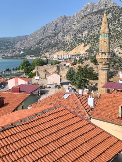 The image captures a panoramic view of the city of Eğirdir, Turkey, with a prominent minaret of a mosque in the foreground, set against a backdrop of a rugged mountain range and a calm body of water, likely Eğirdir Lake. The foreground is dominated by a dense cluster of terracotta-tiled roofs, showcasing the architectural style of the region. Several residential buildings with varying colors and designs are visible amidst the rooftops. Further into the scene, the clear blue water of the lake stretches across the horizon, with a few boats dotting its surface. On the opposite bank, a town with a mix of modern and older buildings is nestled at the foot of the imposing, rocky mountains. The slopes of these mountains are sparsely vegetated, with some areas showing evidence of erosion. The sky is a clear, bright blue, suggesting a sunny day. The overall impression is one of a picturesque and tranquil lakeside city, characterized by its natural beauty and architectural heritage.