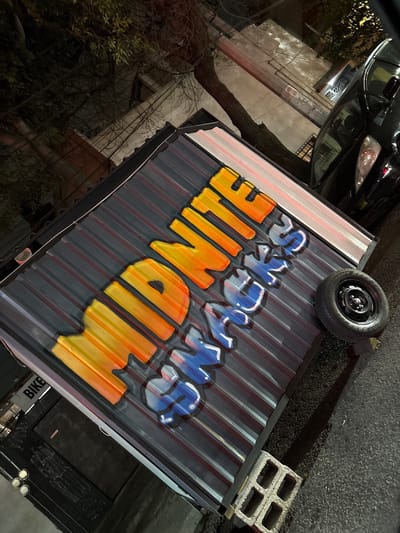 The image shows a low-angle, slightly tilted view of a corrugated metal structure, possibly a food cart or kiosk, with the words "MIDNITE SNACKS" painted on the side in large, colorful graffiti-style lettering. "MIDNITE" is in orange and yellow, while "SNACKS" is in blue and white. The structure is sitting on asphalt pavement, with a spare tire resting on the top left corner and a concrete block on the top right corner. A black car is parked in the background and reflected in the corrugated metal. The surrounding environment appears to be an urban area with trees and buildings. The lighting suggests it is nighttime. The visible letters "BIKE" suggest a possible business connection with bicycles, or it's a sticker. While Tizapan, Mexico is mentioned, there are no obvious visual clues (e.g., signage, distinctive architecture) to confirm the specific location.