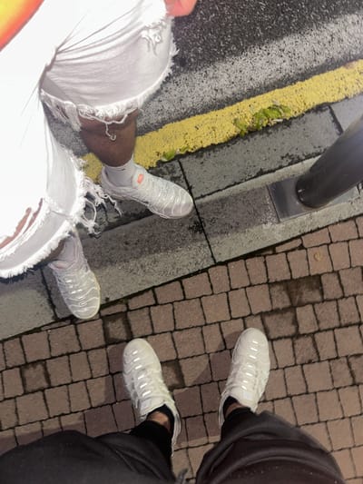 An overhead shot captures the legs and feet of two individuals standing on an urban or semi-urban surface at night. One person, positioned on the left, wears light-colored (likely white) ripped jeans, white socks, and white sneakers. The sneakers are identifiable as Nike Air VaporMax Plus, featuring an orange "TN" logo on the tongue and "VAPORMAX PLUS" text on a side tag. The other person, on the right, wears dark pants and similar white Nike Air VaporMax Plus sneakers with a visible side tag.
The ground comprises multiple textures: dark asphalt with a prominent yellow painted line running vertically, a concrete curb separating the asphalt from brick pavers, and brick pavers on the right side of the image. Small patches of green debris, possibly leafy greens, are visible on and near the yellow line. A black pole, likely a street light or signpost, is anchored to the concrete curb. Several wet spots or small puddles are noticeable on the brick pavers, suggesting recent rain. The scene is illuminated by artificial light, reflecting off the sneakers and wet surfaces, indicating it is nighttime or very low light. The image captures a casual outdoor moment in Dzhebel, Bulgaria.