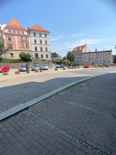 The image captures a view of a street and sidewalk in Świdnica, Poland, on a clear day with a bright blue sky and scattered clouds. In the foreground, a cobblestone sidewalk with white markings indicating parking spaces stretches across the frame. A grassy strip separates the parking area from a road paved with smaller cobblestones.
Further in the background, several cars are parked on the street. To the left, a bright red car is visible. Behind the cars, a low stone wall topped with greenery separates the street level from a slightly elevated area.
Dominating the middle ground and background are several multi-story buildings. On the left, a building with a prominent salmon-pink facade and a red tiled roof features multiple windows. Adjacent to it is a lighter-colored building with a similar red tiled roof. Further to the right, a larger, beige building with numerous windows and a flat roof is visible. Across the street, another building with a red tiled roof and multiple floors is present.
In the distance, a crosswalk with white stripes is visible across the intersection, and a person is walking across it. Traffic lights are also present at the intersection. The environment appears to be urban, with a mix of residential or commercial buildings and public street space. The overall scene suggests a typical European city street on a sunny day.
