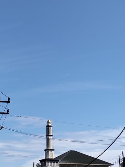 A mosque minaret, topped with a golden dome, rises from the building's green roof against a mostly clear, blue sky. Power lines crisscross the lower portion of the frame, partly obscuring the view. A utility pole stands to the left, further adding to the tangled network of wires. The base of the minaret displays a repetitive arched pattern.