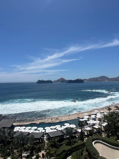 The image depicts a beach scene in Cabo San Lucas, Mexico, taken from an elevated perspective. A prominent cruise ship is visible in the ocean, positioned relatively close to the shore. The beach is lined with white umbrellas and appears to have a moderate number of people present. Waves are breaking along the shoreline. In the foreground, a portion of a resort or hotel is visible, with lush vegetation and landscaped areas. The sky is clear and blue with wispy clouds. Mountains can be seen in the background.