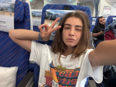 The image shows a woman taking a selfie on an airplane. She is seated in a blue airline seat with travel advertisements printed on the headrest cover. She is winking with one eye and holding up a peace sign with her hand. She is wearing a white t-shirt with a graphic design. Behind her is a male passenger using a cell phone. The airplane interior is visible in the background, including overhead compartments and window curtains. The seat covers advertise travel to the "Great Zhangjiajie" in Chinese. The flight is with China Southern airlines. The city of Baiyun District, China, cannot be confirmed from the image alone.