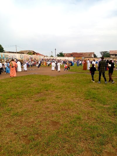 The image shows a large group of people gathered on a grassy field in Beni, Democratic Republic of Congo. The field is located at latitude 0.489 and longitude 29.463. The people are mostly men and boys, some of whom are wearing traditional clothing. There is a large building in the background, with a sign on the wall that reads "BNT ASBLE." The people in the foreground are spread out, some are standing around talking, while others are walking around. Some of the people are holding cell phones. The sky is overcast. The ground is brown with patches of green grass. There are trees in the background.