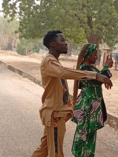 A man and woman are outdoors on what appears to be a college campus. The man, in profile, is wearing a tan two-piece outfit with colorful African-inspired trim. The woman, also in profile, is wearing a green and black dress with a green headscarf. She is holding up a cell phone, seemingly taking a photo or video. They are standing on a paved road with trees and buildings in the background.