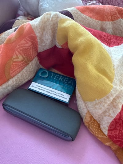 A teal pack of TEREA Turquoise for IQOS ILUMA sticks is placed on a dark gray device case. The pack has Cyrillic text that translates to "This tobacco product is harmful to your health and leads to addiction." The items are on a pink surface, with a colorful patterned blanket draped behind them. The location is the city of Rogosh, Bulgaria.