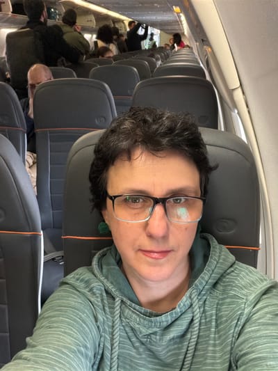 A person with short, dark hair and glasses is sitting in an airplane seat, taking a selfie. They are wearing a green hooded sweatshirt. The airplane cabin is visible behind them, with rows of seats and other passengers. The location is not specified in the image, but the question indicates it is in Guarulhos, Brazil.
