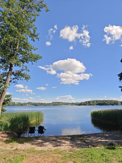This is a landscape photograph of a tranquil lake scene in Būtkiemis II, Lithuania. In the foreground, two empty black camping chairs are set up on a sandy shore, facing the water. To the left of the chairs, a small, dark metal grill sits on the sand. Tall green reeds line the edge of the lake, creating a natural barrier between the shore and the water.

The lake itself is calm, reflecting the bright blue sky above, which is dotted with fluffy white cumulus clouds. The distant shoreline is covered in a dense, dark green forest. The overall setting suggests a peaceful day in nature, likely during the daytime given the bright sunlight and clear sky. The scene evokes a sense of relaxation and serenity, as if awaiting people to sit in the chairs and enjoy the view. There is no visible text in the image.