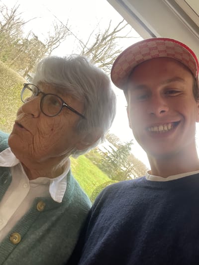 This close-up portrait features two individuals, an elderly woman and a younger male, situated indoors next to a large window revealing an outdoor scene. The location is specified as Chambéry, France.
On the left, an elderly Caucasian woman with short, wavy grey hair wears dark-framed eyeglasses, a white collared shirt, and a light teal/green button-up cardigan with light brown buttons. Her face exhibits prominent wrinkles, and she is looking upwards and to her left, with her mouth slightly ajar.
To the right, a younger Caucasian male, appearing to be in his twenties, is present. He wears a red and beige baseball cap adorned with a repeating "G" logo pattern. His short brown hair is visible beneath the cap. He is smiling directly at the camera, revealing his upper and lower teeth. He wears a dark blue crew-neck sweater over a white collared shirt, the collar visible at his neckline.
The background, visible through the window, depicts a bright outdoor environment. Bare deciduous trees are prominent, suggesting late winter or early spring. A dense green hedge is visible, along with patches of vibrant green grass and some yellow foliage or flowers. The bright light suggests daytime. The composition indicates a selfie or close-up photograph taken by the male individual, capturing both subjects in an intimate setting.