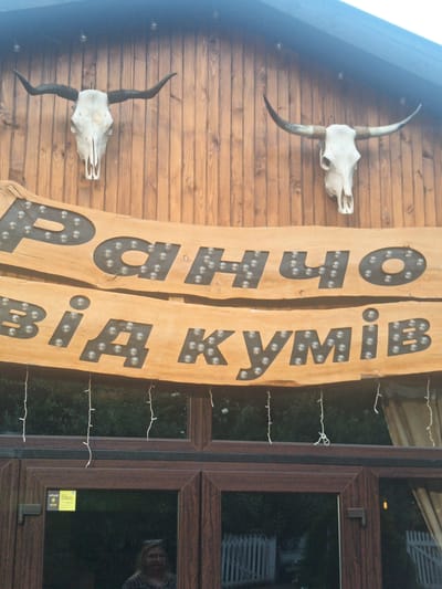 The image shows the exterior of a building with a wooden facade. Two animal skulls with large horns are mounted on the wall, one on the left and one on the right. A large wooden sign with carved letters, illuminated by small bulbs, is mounted below the skulls. The text on the sign appears to be in Cyrillic script. Below the sign, there is a dark-framed window with what looks like a reflection of a person. String lights hang from the roof above the window. The overall impression is of a rustic or themed establishment. The context of "Tyihlash, Ukraine" suggests this is likely a business or a landmark in that specific location.