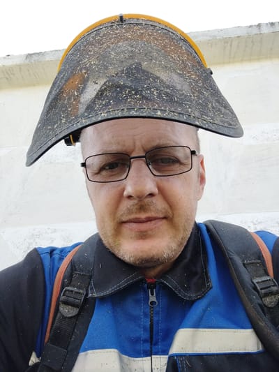A middle-aged Caucasian man wearing glasses and a protective face shield made of mesh and plastic is shown from the chest up. The man is wearing a blue and black work uniform with orange straps from a backpack visible over his shoulders. The face shield is dirty with debris, suggesting it has been used for outdoor work. The background appears to be a light-colored, textured wall. The image was taken outdoors in a location that is outdoors in the city of Гожскі сельскі Савет, Belarus.