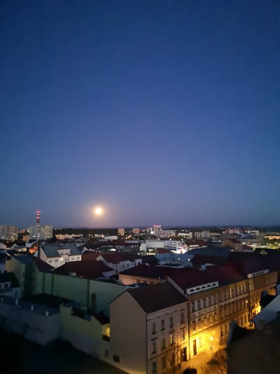 The image presents an elevated night-time view of an urban landscape, likely Pardubice, Czechia. The scene is dominated by a deep, clear blue sky, indicating late twilight or night. A bright, full or near-full moon is prominently visible on the left side of the sky, casting a soft, ethereal glow.
Below the expansive sky, the city sprawls with a dense array of buildings. Many structures feature traditional European architecture, characterized by pitched roofs, some with warm red tiles and others in darker tones. Numerous windows within these buildings are illuminated, creating scattered points of warm light against the shadowed facades. Streetlights below cast a warm, yellow glow on the streets and lower sections of the buildings, highlighting their architectural details, particularly visible in the ornate facades of buildings along a street in the foreground-right.
A tall, red and white striped industrial chimney or smokestack stands out on the left side of the frame, an industrial landmark amidst the residential and commercial structures. In the far distance, a faint line of city lights stretches towards the horizon, suggesting the city's vastness.
No people are visible in the frame, and there is no discernible activity taking place, lending a calm and tranquil atmosphere to the illuminated cityscape. The weather appears clear with no clouds. No visible text can be deciphered on any of the buildings or signs. The overall impression is one of a peaceful urban night scene.