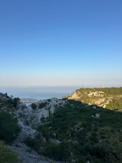 The image depicts a mountainous coastal landscape under a clear blue sky. In the foreground, a rocky slope covered with sparse green vegetation descends into a valley. Beyond this, a body of water, likely the Adriatic Sea, is visible with a distant coastline shrouded in a light haze. To the right, a rocky ridge is densely forested with pine trees, and a telecommunications tower stands on its summit. The scene suggests an elevated viewpoint looking out over the sea and surrounding terrain. The location is identified as Brajici, Montenegro.
