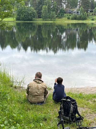 In a natural outdoor setting by a lake, two individuals are the subjects. One person, wearing a dark blue long-sleeved shirt, is seated facing the water, with their head down as if looking at something. Beside them, a gray and black backpack rests on the grassy bank. The second individual, dressed in a camouflage hoodie, is also seated by the water, facing away from the camera. Their head is down, suggesting a similar focus on the lake.
The scene is characterized by the calm surface of the lake, which reflects the trees and cloudy sky, creating a mirrored effect of the surrounding landscape. The shoreline is covered in green grass, with some patches of sand visible. In the distance, across the lake, a line of trees and what appear to be houses or buildings can be seen, hinting at a populated area nearby. The overall atmosphere suggests a peaceful, leisurely activity, possibly fishing or simply observing nature. The lighting indicates it is daytime, with overcast weather, as the sky appears bright but diffused. There is no visible text in the image. The location context provided places this scene in Mars, Russia.