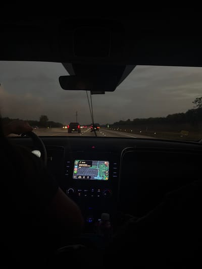 The image displays a forensic perspective from inside a vehicle, looking forward through the windshield. The scene depicts a highway environment during twilight or nighttime, characterized by an overcast sky and wet road surfaces, indicative of recent or ongoing precipitation.
**People:**
* One individual, the driver, is partially visible on the left, with their left hand gripping the steering wheel. Their upper body is largely obscured by shadow.
* Another individual, presumed to be a front passenger, has their lower right leg and foot visible in the lower right quadrant. They are holding a dark electronic device, possibly a mobile phone or game controller.
**Subjects/Objects (Interior):**
* The driver's side features a steering wheel.
* The central dashboard contains an illuminated infotainment screen displaying a navigation application. The map clearly shows "Boca Raton, FL," and street names such as "Military Trail Rd" and "NW 2nd Ave" are visible. A destination or point of interest labeled "Southern Blvd" is indicated as 10 miles away.
* Below the infotainment screen are illuminated climate control buttons and dials.
* A white-capped bottle, possibly containing a beverage, is visible resting in a console compartment or cup holder.
* A rearview mirror is suspended from the vehicle's ceiling, slightly obscuring the upper central view.
**Subjects/Objects (Exterior):**
* The vehicle is traveling on a multi-lane highway with wet asphalt, reflecting lights.
* Multiple vehicles are visible ahead, displaying illuminated red taillights.
* Road reflectors or delineators line the shoulders of the highway.
* Dark, indistinct vegetation (trees and bushes) borders the right side of the highway.
* The sky overhead is uniformly overcast and dark gray.
**Actions/Interactions:**
* The vehicle is in motion on the highway.
* The driver is operating the vehicle.
* The navigation system is actively displaying a route.
* The passenger is occupying themselves with an electronic device.
**Location:**
The location is positively identified as Boca Raton, Florida, United States, based on the visible labels and map context provided by the in-car navigation system. The navigation indicates proximity to "Military Trail Rd" and "NW 2nd Ave" within Boca Raton, with "Southern Blvd" as a distant point.