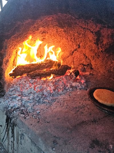 A wood fire burns intensely inside a rustic oven or pit. Flames lick around a dark, charred log at the center, transitioning to glowing embers on the right. The oven walls are textured with a sandy, reddish-brown buildup. A circular vent with a cork rests near the top. Traces of dark, dripping residue stain the bottom, suggesting past fires and high temperatures. The fire's glow illuminates the gritty texture and intense heat.