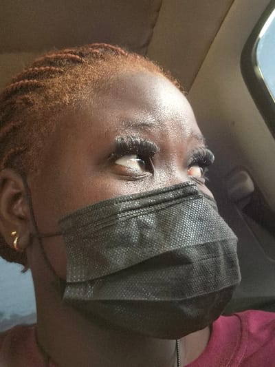 A low-angle shot features a Black person wearing a black face mask. The mask covers the nose and mouth, obscuring the lower half of the face. Dark eyelashes are visible above the mask, and the skin appears smooth. The person has reddish-brown hair. One gold earring is visible. The background suggests an interior space, possibly a car, with a window and a beige seat.