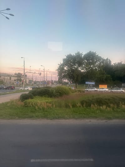The image depicts a street scene in Lublin, Poland, likely at dusk given the soft lighting and pastel sky. In the foreground, a two-lane asphalt road is visible, with a white line marking a lane divider. To the left and center of the frame, a grassy median separates the road from a sidewalk and a dense line of trees and bushes.

Several cars are present on the road, appearing to be stopped or moving slowly at an intersection. Traffic lights are visible, with one showing red. Beyond the road, a modern building complex with white and grey facades is seen, along with streetlights and overhead wires, suggesting an urban environment. A blue street sign with white lettering reads "Al. Tysiąclecia", indicating the name of the avenue. In the background, more trees and foliage are present, and a construction site with a yellow sign that reads "UWAGA!" (Attention!) is visible. A few pedestrians can be observed walking near the intersection and along the sidewalk. The overall impression is a typical urban street scene in Poland.