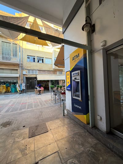 The image shows a street scene in Nicosia, Cyprus. In the foreground, there is an ATM machine with "ATM cash 24h" displayed on it, as well as the "Euronet" logo. The ATM is located next to a building entrance. The street is paved with stone tiles and there are red lines marking a pedestrian crossing. In the background, there are shops and cafes with outdoor seating. One shop has clothing displayed on racks outside. People are seated at tables outside a cafe, engaged in conversation. The buildings are typical of Mediterranean architecture, with balconies and shutters. The scene is illuminated by sunlight, casting shadows on the ground.