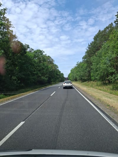 A view from inside a vehicle traveling on a two-lane asphalt road, lined with dense green trees on both sides. The sky above is blue with scattered white clouds. In the distance, a white car with its trunk open is visible, and another vehicle is behind it. The road has solid white lines marking the edges and a broken white line dividing the lanes. The area appears to be a rural or semi-rural location, consistent with the location of Przyłubie, Poland. The scene depicts a typical daytime drive through a forested area.