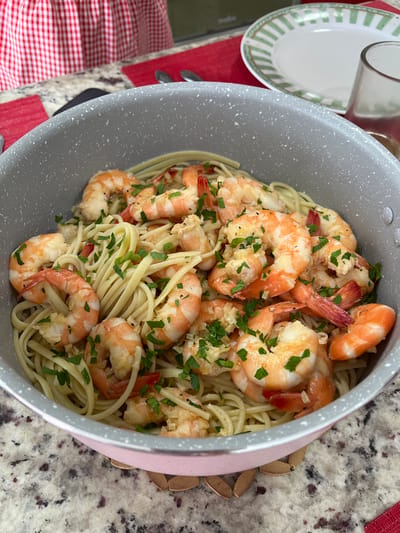 This close-up image showcases a large, grey speckled pot filled with a generous serving of shrimp scampi or garlic butter shrimp and linguine pasta. The plump, pinkish-orange shrimp are interspersed throughout the light-colored pasta, all glistening with what appears to be a buttery garlic sauce. Freshly chopped green parsley is sprinkled abundantly over the dish, providing a vibrant contrast.
The pot has a light pink or rose gold colored exterior rim visible on the right side. The dish is placed on a speckled countertop, likely granite or quartz, featuring a mix of white, grey, and reddish-brown tones. In the upper left corner, a white plate with a green striped pattern around its edge is partially visible, alongside a clear glass or cup. A red and white checkered cloth or placemat can be seen on the bottom left. Additionally, a few dried, brown, claw-like objects are visible on the countertop behind the pot on the right, possibly decorative items.
The scene appears to be set in a home, possibly a kitchen or dining area, illuminated by bright, even lighting, suggesting daytime. The meal looks freshly prepared and appetizing, evoking a sense of warmth and domestic comfort. No visible text is present in the image. Although the context provided indicates the location is Barrio de Nuevo León, Mexico, there are no specific visual cues within the photograph itself to confirm this geographical detail.