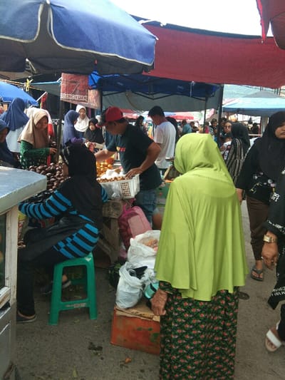 This image captures a lively outdoor market scene in Baleendah, Indonesia, under the shade of various awnings and tarps, primarily blue and red. The setting is bustling, filled with people engaged in commerce.

In the foreground, to the left, a person wearing a black hijab and a blue and black striped long-sleeved top is seated on a bright green plastic stool, attending a stall with dark, round fruits, likely salak. On the right, a woman seen from behind wears a vibrant lime green hijab and a dark green floral-patterned long skirt, standing and observing the market. Her left hand, appearing somewhat aged, is visible. White plastic bags filled with goods and an orange crate are on the ground near her.

In the midground, a man in a red cap and a black t-shirt is actively working at a stall, arranging or serving light-colored fruits, possibly rambutan or longan, from a white plastic basket. To his left, several women, mostly wearing hijabs, are browsing or purchasing items from stalls. One woman with a light brown hijab and green patterned top is reaching for fruit. To the right, more market-goers, including women with children, move through the busy aisles.

The scene depicts a typical market environment with vendors and customers. The lighting suggests it is daytime. A red sign is partially visible under a blue awning, with legible text that appears to read "ODENG KUAH." The overall atmosphere is busy and functional, characteristic of a local market.