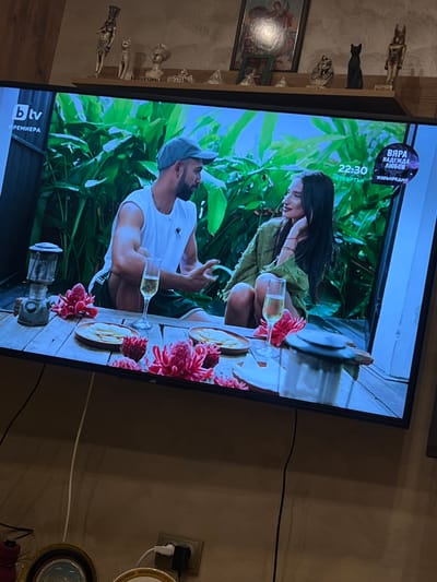 A television displays a scene depicting a man and a woman seated at a table with food and drinks, amidst lush greenery. The TV rests on a wooden surface where decorative figurines are placed. The setting is presumably indoors in Popintsi, Bulgaria. A wall outlet with plugs and decorative plates are also visible in the background.