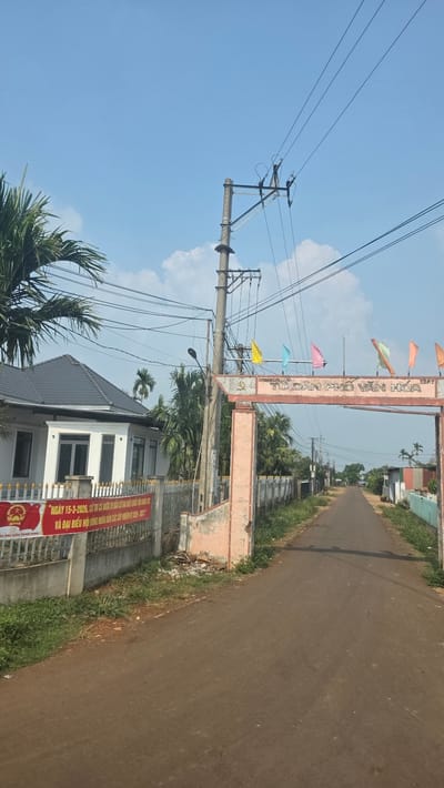 The image depicts a street scene in Xuân Long, Vietnam. A road stretches into the distance, bordered by vegetation and structures. An ornate archway with colorful flags spans the road, bearing text in Vietnamese. To the left, a utility pole with power lines stands prominently. A two-story building with a gray tiled roof and white walls is visible. A banner with red and gold colors, also with Vietnamese text, is attached to the building's fence. The sky is blue with scattered clouds, and palm trees are visible in the foreground.