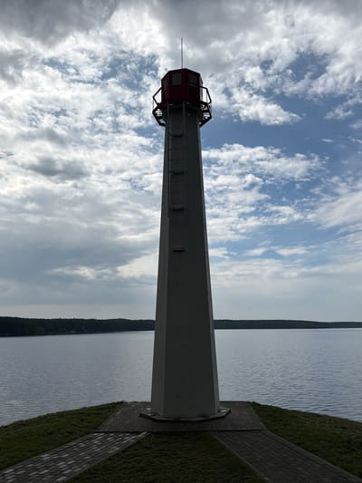 A tall, white lighthouse stands on a grassy knoll overlooking a calm body of water. The lighthouse has a red top with glass windows and a metal railing. A ladder is attached to the side of the lighthouse. The water stretches out to a tree-lined horizon under a cloudy sky with patches of blue. Paved pathways lead away from the base of the lighthouse across the grass. This lighthouse is located in Zarechny, Russia.