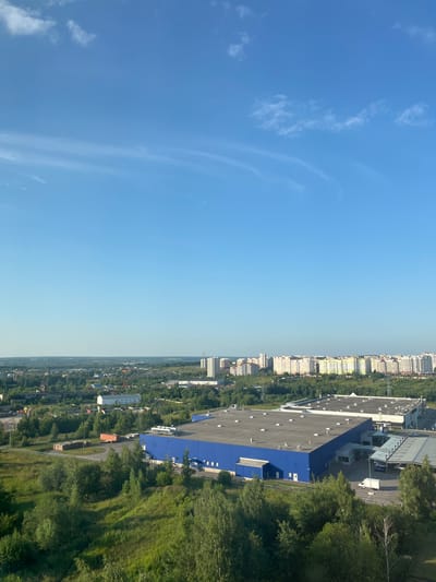 This is a high-angle, rotated photograph of a suburban landscape under a clear blue sky with wispy clouds. The scene is divided between the vast expanse of the sky on the left and a densely populated urban area on the right.
The urban area consists of numerous residential buildings, appearing to be apartment blocks, with light-colored facades and multiple stories. These buildings are interspersed with a significant amount of greenery, indicating a treed environment within the city. In the foreground of the urban area, a large, modern industrial building with a bright blue facade and a dark roof is prominent. This building has many small white squares visible on its blue side, which may be windows or decorative elements. The roof is flat and appears to have numerous evenly spaced circular fixtures, possibly skylights or vents. The industrial building is surrounded by trees and appears to be situated near a road or pathway.
The time of day is suggested to be daytime, likely late morning or early afternoon, given the bright sunlight and clear sky. The weather appears to be fair and sunny. The location is identified as Oryol, Russia. No specific activity or event is depicted, and no people are clearly visible. The overall impression is one of a developing or established urban area with significant green spaces.