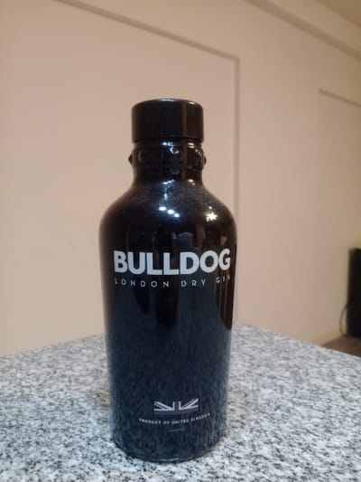 A dark, cylindrical glass bottle of Bulldog London Dry Gin stands vertically on a speckled grey and white granite surface. The bottle's label, in white capital letters, displays "BULLDOG" prominently, with "LONDON DRY GIN" positioned beneath it. Near the bottom, a white stylized Union Jack flag is depicted, accompanied by the text "PRODUCT OF UNITED KINGDOM." The bottle is sealed with a black screw cap. The background consists of a plain, light beige wall featuring subtle recessed paneling. A dark, indistinct object is partially visible in the far right background. The scene is static and illuminated from the front, creating reflections on the bottle's surface. This image is situated in Lomas de Zamora, Argentina.