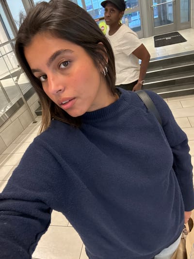 The image depicts an indoor scene within the Fashion District shopping mall in Philadelphia, Pennsylvania, United States. In the immediate foreground, a light-skinned female with dark brown hair, wearing a navy blue knitted sweater and multiple silver hoop earrings, takes a selfie, looking directly into the camera. In the mid-ground, a dark-skinned female, wearing a black baseball cap, a white t-shirt, large yellow hoop earrings, and a smartwatch on her left wrist, stands near a glass entrance. She is holding a light brown paper bag in her left hand. A doormat bearing the text "FASHION DISTRICT" is visible on the floor inside the glass doors. A light-colored tiled floor extends through the foreground and mid-ground, leading to stairs or a ramp near the entrance. No direct interaction between the two individuals is evident.