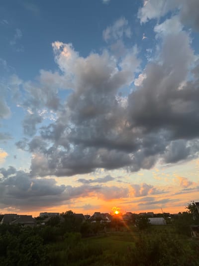 A vertical, high-angle shot captures a sunset over a rural residential area, possibly in Slobozia, Moldova. The sky is a canvas of dramatic clouds, ranging from dark gray masses to lighter, sun-kissed formations tinged with orange and yellow. The setting sun, a bright orange disc, is partially obscured by clouds and trees, casting a warm glow across the horizon. Below the sky, a dense canopy of green trees and vegetation forms the foreground. Scattered amongst the trees are the rooftops of houses, with some structures appearing to be of older construction and others more modern, including a greenhouse with a white covering. The overall impression is one of a serene, end-of-day scene in a tranquil, semi-rural setting.
