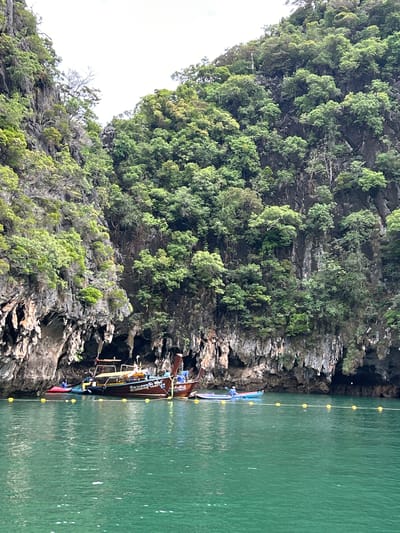 The image captures a serene view of Phang Nga Bay, near Khlong Khian, Thailand. The scene is dominated by towering limestone cliffs covered in lush green vegetation, with the emerald green water of the bay gently lapping at the shore. Several long-tail boats, characteristic of Thai waterways, are anchored in the water. Two of these boats are brown, one of which has a white canopy and is adorned with colorful ribbons. There are also two kayaks, one red and one blue, floating near the boats. A rope with yellow buoys stretches across the water, marking a boundary. A few individuals are visible on the boats and in the kayaks, suggesting a tourist or recreational activity. The sky above is overcast, casting a soft light on the entire scene. The overall impression is one of natural beauty and tranquility.