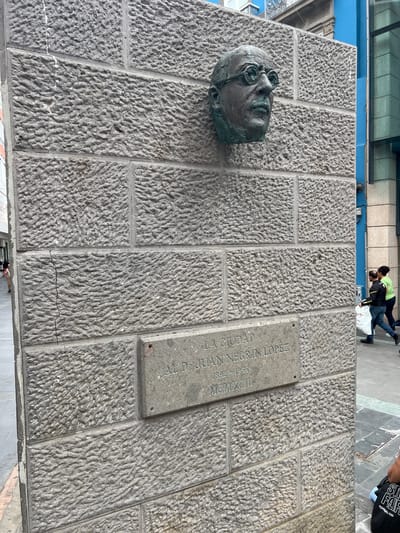 The image shows a stone wall in Las Palmas De Gran Canaria, Spain. A bronze head is mounted on the left side of the wall. To the right of the head, a plaque reads "LA CIUDAD AL Dr. JUAN NEGRIN LOPEZ 1892-1956 MCMXCIII". Two people are visible in the background. One is wearing a green jacket. A third person is partially visible in the upper right corner.