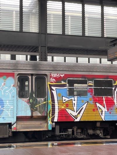 The image shows a graffiti-covered train car at a train station in Aveiro, Portugal. The train is red and yellow with blue and white graffiti art on its side. The graffiti art includes stylized letters and shapes. The train car has two doors and several windows. The station has a modern architectural design with a metal roof and large windows. The platform is made of concrete and has yellow and black lines marking the edge. There are no people visible in the image. The scene appears to be during the daytime, with overcast lighting conditions. The overall impression is one of urban decay and artistic expression.