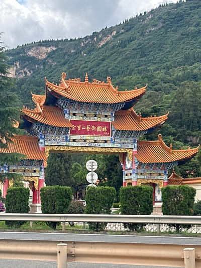 A traditional Chinese archway stands in Xishan District, China. The archway features multiple tiers of orange-tiled roofs with ornate, colorful detailing in blue and gold. A red plaque with Chinese characters is mounted above the central arch. The archway is surrounded by lush green trees and bushes, and a mountainous, forested landscape forms the backdrop under a cloudy sky. A road with a metal guardrail is visible in the foreground.