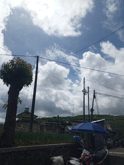 This daytime photograph captures a vibrant outdoor scene in what appears to be a rural or semi-rural setting in Hinabangan, Philippines. The sky dominates the upper portion of the image, showcasing a striking blue expanse dotted with numerous white, fluffy cumulus clouds, suggesting partly sunny weather.

In the foreground, a sturdy, dark-barked tree with dense green foliage stands prominently on the left. Below it, a low stone wall supports a patch of vibrant green plants, possibly crops. Several utility poles with a complex web of overhead electrical or communication wires crisscross the frame, adding a layer of infrastructure to the landscape.

Beyond the foreground elements, a modest building resembling a mosque or musala is visible, featuring a distinct dome and a yellowish-tiled roof, with loudspeakers mounted near its peak. Further to the right, a blue umbrella provides shade, likely covering a small vendor's stall, beneath which several large plastic water bottles are placed. A red motorcycle, partially obscured, is parked nearby, with its license plate clearly visible, displaying "PL 8297 NL". Bags, possibly containing goods, rest on the ground next to the motorcycle.

The background reveals sloping green hills, indicative of agricultural land or natural terrain, with structures and scattered vegetation. A large metal lattice power transmission tower is discernible on one of the distant hillsides.

No people are visible in the image, but the presence of the motorcycle, umbrella, and water bottles suggests ongoing daily life and possibly commercial activity within the community. The overall atmosphere is one of everyday tranquility, with nature and human habitation coexisting under a bright sky.