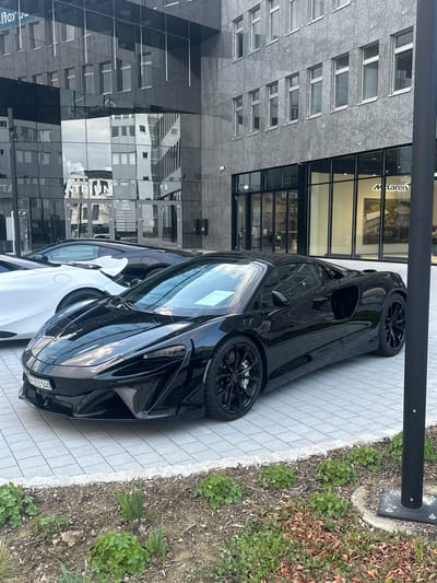 The image captures a black McLaren sports car parked on a paved area next to a modern building in Glattbrugg, Switzerland. The building's facade features a combination of grey stone and reflective glass. "McLaren" signage is visible on the building's window. A white car is partially visible next to the McLaren. A black pole stands near the edge of the pavement. Adjacent to the pavement is a patch of ground with vegetation and loose leaves. The license plate of the black McLaren is visible.