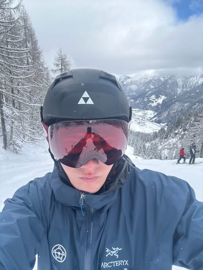 A male individual is depicted taking a selfie on a snow-covered ski slope, positioned above the town of Bad Hofgastein, Austria. The subject wears a black ski helmet with a white Fischer logo, red mirrored goggles that reflect a person holding a phone, and a dark blue Arc'teryx Gore-Tex jacket featuring a circular turtle-like emblem on the left chest. The immediate environment consists of a groomed ski piste, flanked on the left by snow-covered deciduous conifer trees. In the mid-ground to the right, two other skiers are visible on the slope. The background reveals a valley containing buildings and infrastructure consistent with the town of Bad Hofgastein, nestled among snow-covered forests and distant rugged mountain peaks partially obscured by an overcast sky.