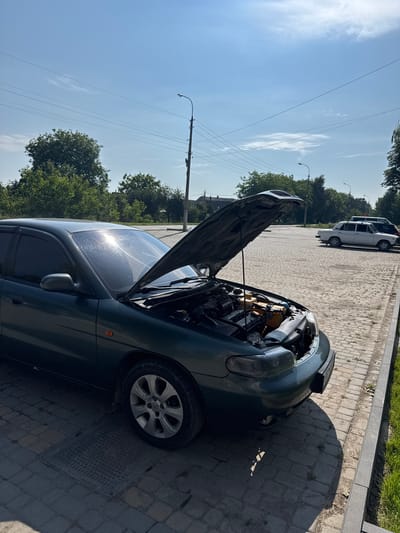 The image depicts a dark green car with its hood open, parked on a cobblestone surface in Chemerivtsi, Ukraine. The car appears to be a sedan, possibly undergoing maintenance. The scene is outdoors under a clear blue sky with scattered white clouds. A lamppost is visible in the background, along with trees and other parked vehicles. The sun is casting a strong glare on the car's windshield. The ground is made of interlocking stone pavers, and there is a strip of green grass to the right of the frame.