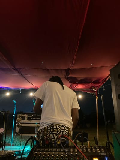 An individual is depicted from behind, operating audio equipment at an outdoor nighttime event in Murera ward, Kenya. The person wears a white short-sleeved t-shirt and patterned trousers with black and white geometric designs; their dark hair is styled in a single braid or dreadlock tail. In the immediate foreground, a Yamaha sound mixing console with numerous controls and attached red, blue, and black cables is visible. Behind the individual, two stacked black equipment cases, one bearing a "Serato" logo, are positioned. To the left, a partial drum kit, including a silver drum and a cymbal, is present. Two microphone stands with microphones are situated further back. A large, dark speaker cabinet is partially visible on the far right. The scene is set under a dark red fabric canopy supported by visible framework. String lights with globe bulbs and colored stage lighting (blue and purple hues) illuminate the area. The ground is grass, and the distant background shows a dark night sky with faint, indistinct figures of other attendees.