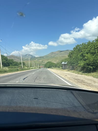 The image, taken from the interior of a vehicle, shows a view of a rural road in Zangakatun, Armenia, on a clear, sunny day. The road stretches into the distance, flanked by green trees and rolling hills. Power lines run along the left side of the road, supported by utility poles. A small, rustic wooden structure is visible on the right side of the road, partially obscured by trees. The dashboard of the car, including the Mercedes-Benz emblem, is visible in the foreground. The sky is blue with scattered white clouds, and the overall scene depicts a peaceful, scenic drive through the Armenian countryside.