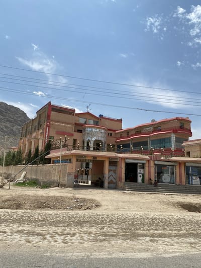 The image depicts a building in Dand, Afghanistan. The building has multiple stories, is peach-colored with red trim, and features a series of arched windows and balconies. There are smaller businesses located in the front of the building. The ground in front of the building is dirt and appears cracked. Mountains are visible in the background on the left side. Several people are seated or standing in front of the businesses. Power lines stretch across the upper portion of the image. The sky is blue with some scattered clouds.