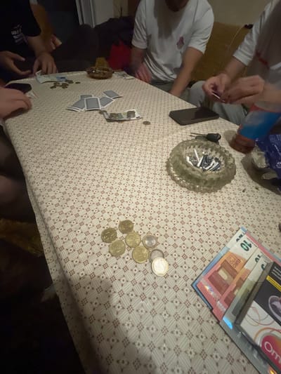 The image depicts a group of people around a table covered with a patterned tablecloth. The table holds assorted items including Euro banknotes, Euro coins, playing cards, a smartphone, a car key, an ashtray filled with cigarette butts, and a pack of cigarettes. Two individuals are visible, one wearing a white t-shirt and another in a dark shirt. The location cannot be determined as Krumovgrad, Bulgaria. No specific building or landmark details are visible to identify it, only the items on the table and the people.